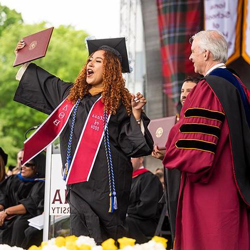 A student holds her diploma and cheers on stage.